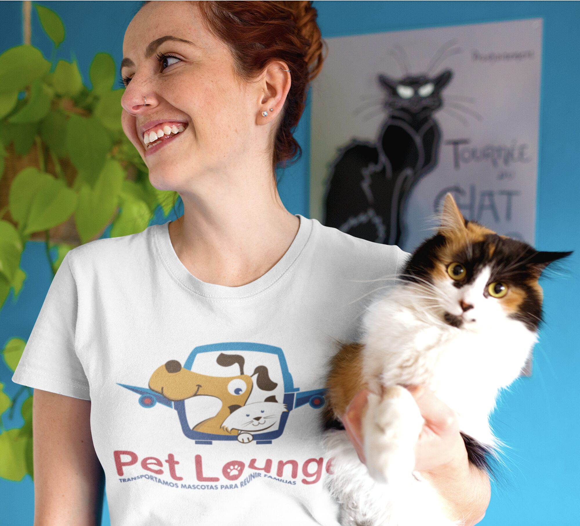 A smiling woman in a "Pet Lounge" T-shirt holding a fluffy calico cat in front of a blue wall with a cat poster.